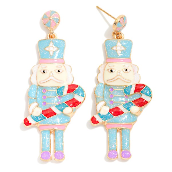 Candy Cane Nutcracker Drop Earrings featuring a whimsical glitter enamel nutcracker holding an oversized candy cane, hanging from a peppermint swirl post. Sweet details and a shimmering finish give this holiday classic a fresh, playful update. A must have for Nutcracker ballet season, dance moms, and candy themed Christmas parties. Trendy nutcracker designs sell quickly every holiday season and make memorable gifts.