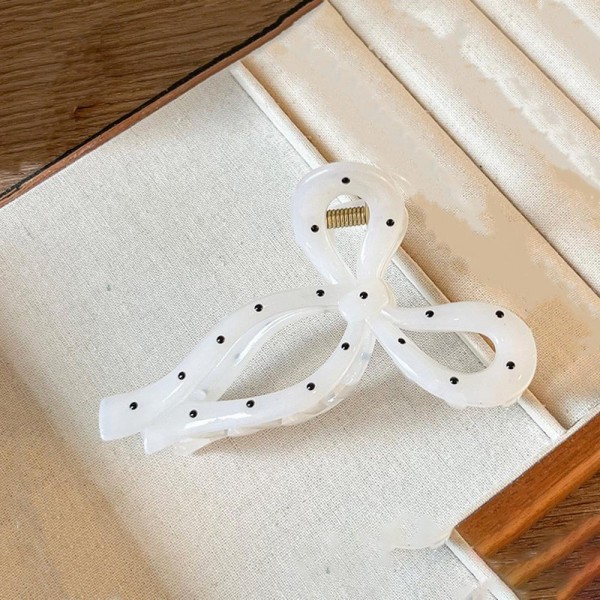 Bow shaped hair claw clip in a glossy acrylic finish with dotted detailing across the loops and a firm hinge that holds a twist or a half up style all day. The open bow silhouette reads delicate from the front while the teeth do real work underneath. Light enough that it never drags on fine hair. Bow claw clips are one of the fastest turning hair accessories on a peg wall right now.