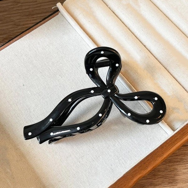 Bow shaped hair claw clip in a glossy acrylic finish with dotted detailing across the loops and a firm hinge that holds a twist or a half up style all day. The open bow silhouette reads delicate from the front while the teeth do real work underneath. Light enough that it never drags on fine hair. Bow claw clips are one of the fastest turning hair accessories on a peg wall right now.