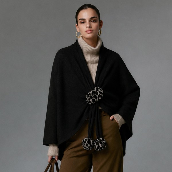 Knit cape wrap with faux fur pom ties at the front, cut wide enough to drape over the shoulders and fall past the hip. The soft brushed yarn feels substantial without weighing down a shoulder, and the open silhouette layers over almost anything on the rack. Sizing is forgiving, so one piece works across a wide customer range. A strong window piece that drives a higher ticket through fall and holiday.