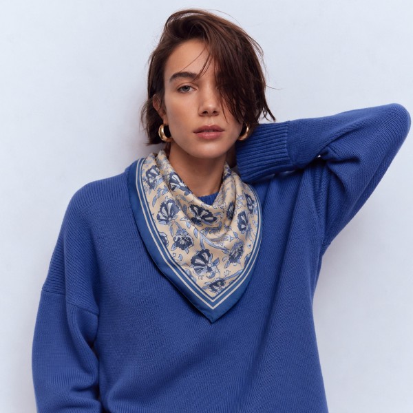 Ribbon and floral print square scarf in a fluid satin-feel woven, finished with a fine contrast border. Oversized enough to knot at the waist as a sarong-style wrap, drape across the shoulders, or fold into a neck scarf. The bow-and-botanical motif lands squarely on the ribbon trend boutiques are chasing this season. One buy, three ways to wear it, which makes it an easy sell on the floor.