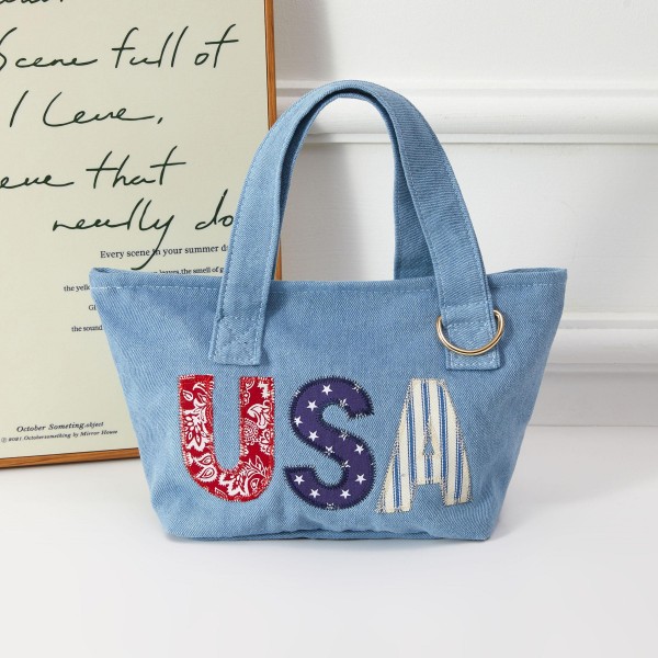A patriotic denim mini tote featuring bold 'USA' applique letters in stars-and-stripes fabric, finished with a metal ring accent. Sturdy construction and comfortable top handles make it practical for everyday use while delivering strong seasonal appeal around summer holidays. A natural add-on for boutiques building Fourth of July and Memorial Day merchandise displays, it appeals to patriotic shoppers and functions equally well as a gift item or impulse buy.