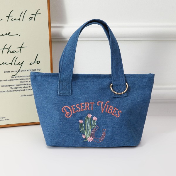 A boho-chic canvas mini tote featuring a hand-embroidered 'Desert Vibes' script and cactus motif, finished with a metal ring accent for added edge. The denim-look fabric gives this bag a laid-back, festival-ready aesthetic that resonates with free-spirited shoppers. Lightweight and roomy for everyday carry, it works as a beach bag, farmers market tote, or casual go-to. A strong seller for boutiques targeting lifestyle and festival fashion customers.