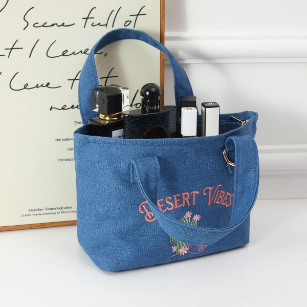 A boho-chic canvas mini tote featuring a hand-embroidered 'Desert Vibes' script and cactus motif, finished with a metal ring accent for added edge. The denim-look fabric gives this bag a laid-back, festival-ready aesthetic that resonates with free-spirited shoppers. Lightweight and roomy for everyday carry, it works as a beach bag, farmers market tote, or casual go-to. A strong seller for boutiques targeting lifestyle and festival fashion customers.